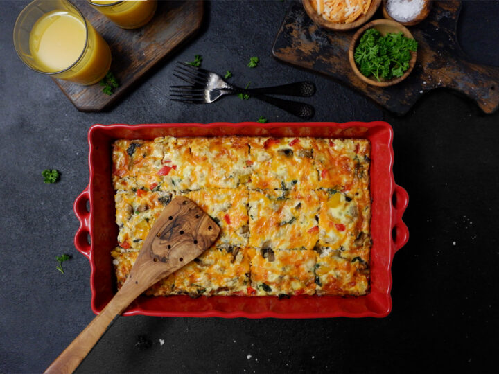 breakfast casserole in a red casserole dish with a wooden spoon on top. Top left 2 glasses of OJ on a wood board, top right parsley, shredded cheese and coarse salt in small wooden bowls on top of a small wooden board