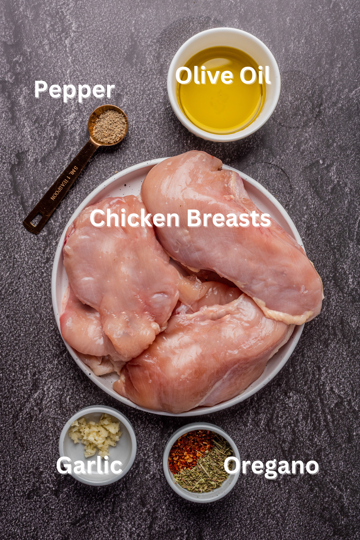 3 raw chicken breasts on a plate in the middle with the marinade ingredients of pepper on a wood spoon top left, top right in a small white bowl olive oil, bottom right in a small bowl chili flakes and oregano.