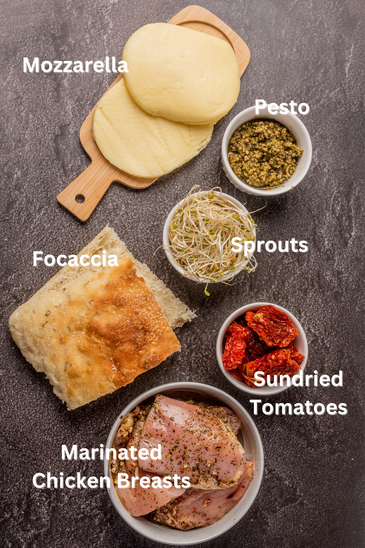 Sandwich ingredients. Top left on a small wood board mozzarella slices, to the right in small white bowls starting at top right pesto, alfalfa sprouts, sundried tomatoes, to the left of that focaccia slices and bottom is the marinated chicken breasts.
