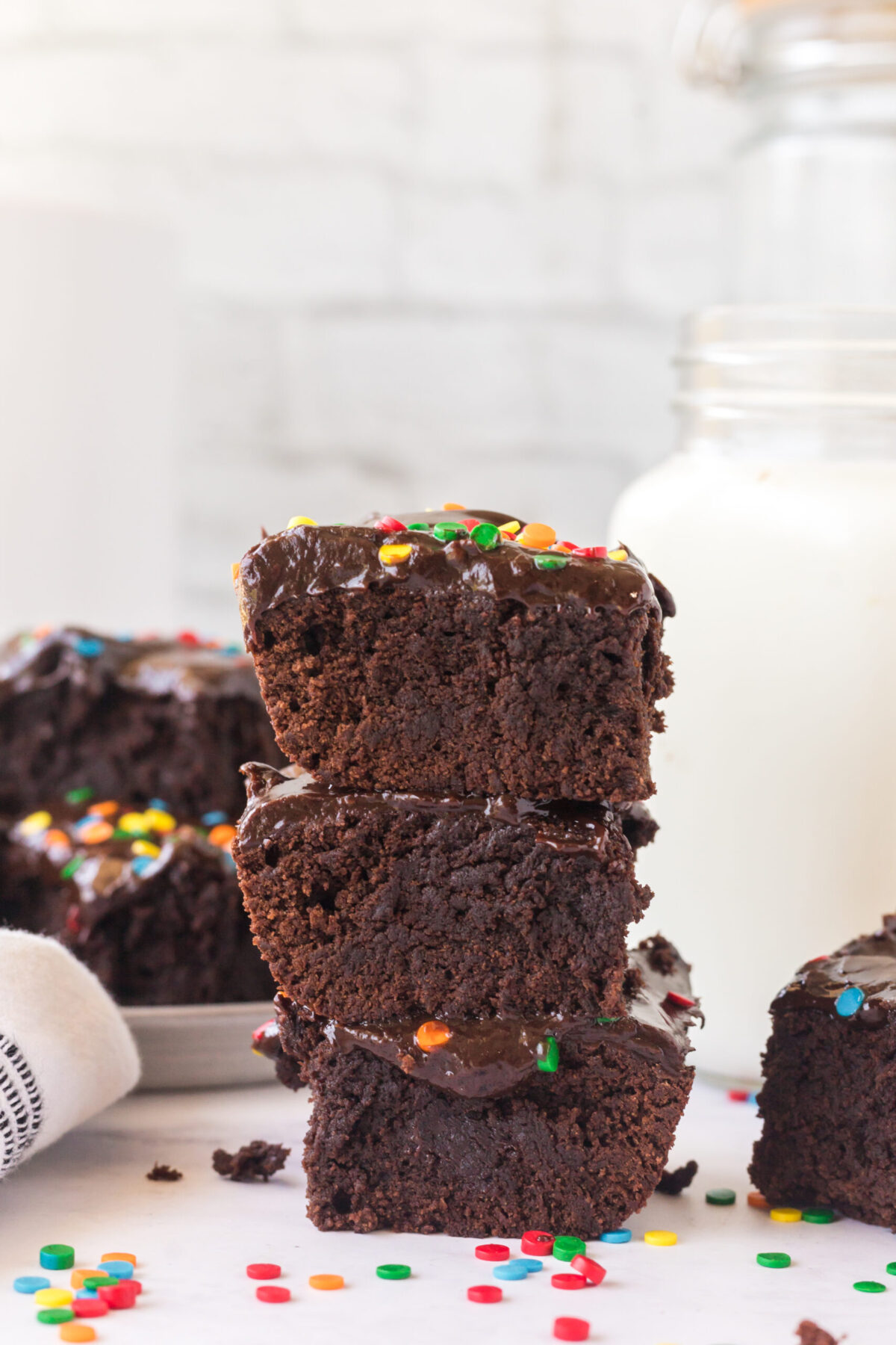 stack of 3 cosmic brownies recipe in the middle, milk jug back right, brownies on a plate back left. rainbow sprinkles on surface. white brick background, white surface.