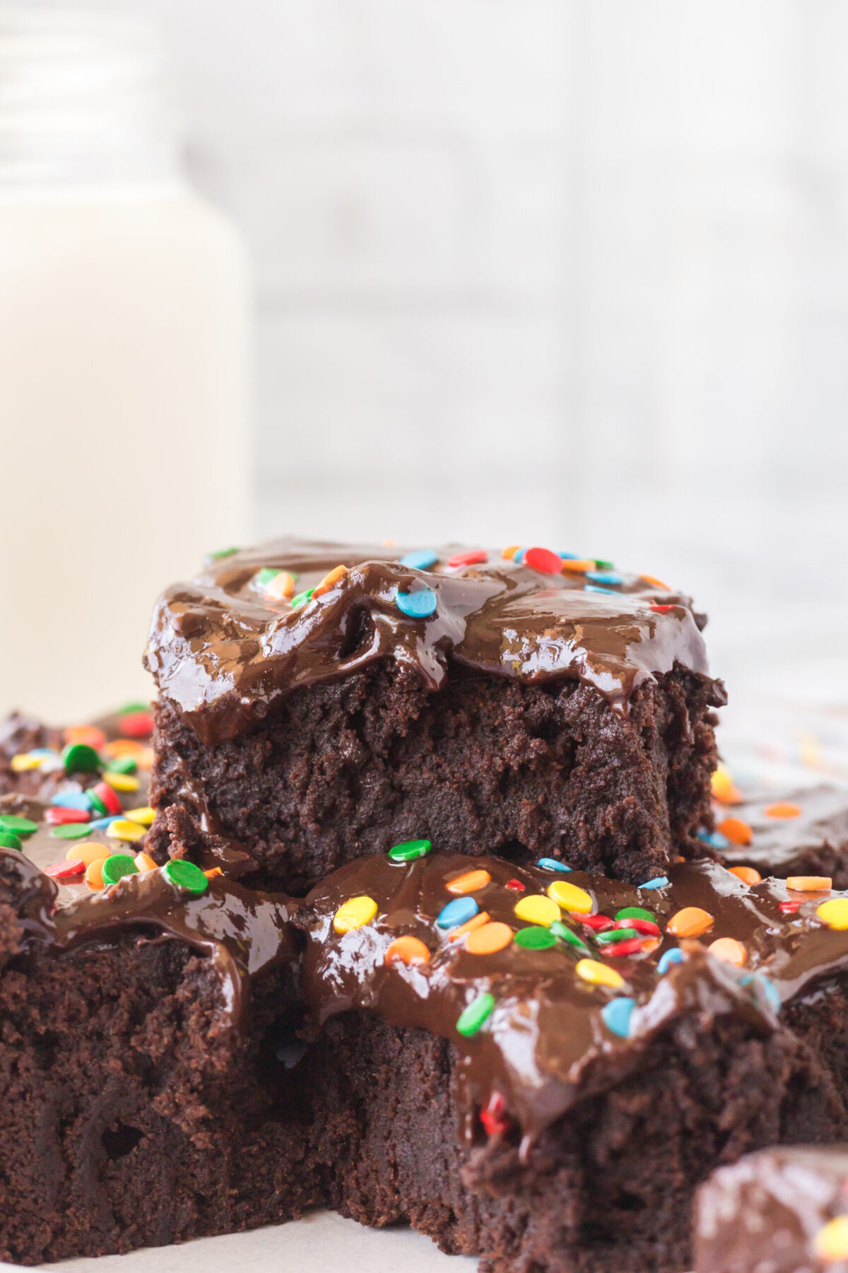 1 brownie with a bite out of it sitting on top of 2 other brownies. milk jug back left, white brick background, white marble surface