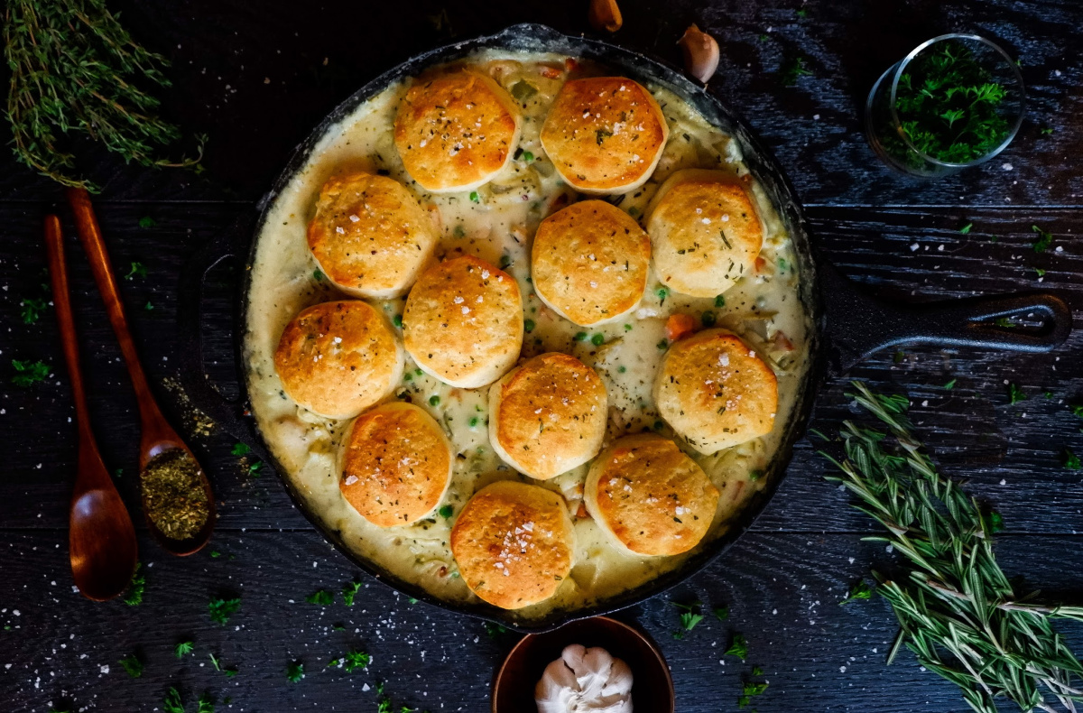cast iron chicken pot pie with biscuit topping on black table with herbs, garlic, and two wooden spoons