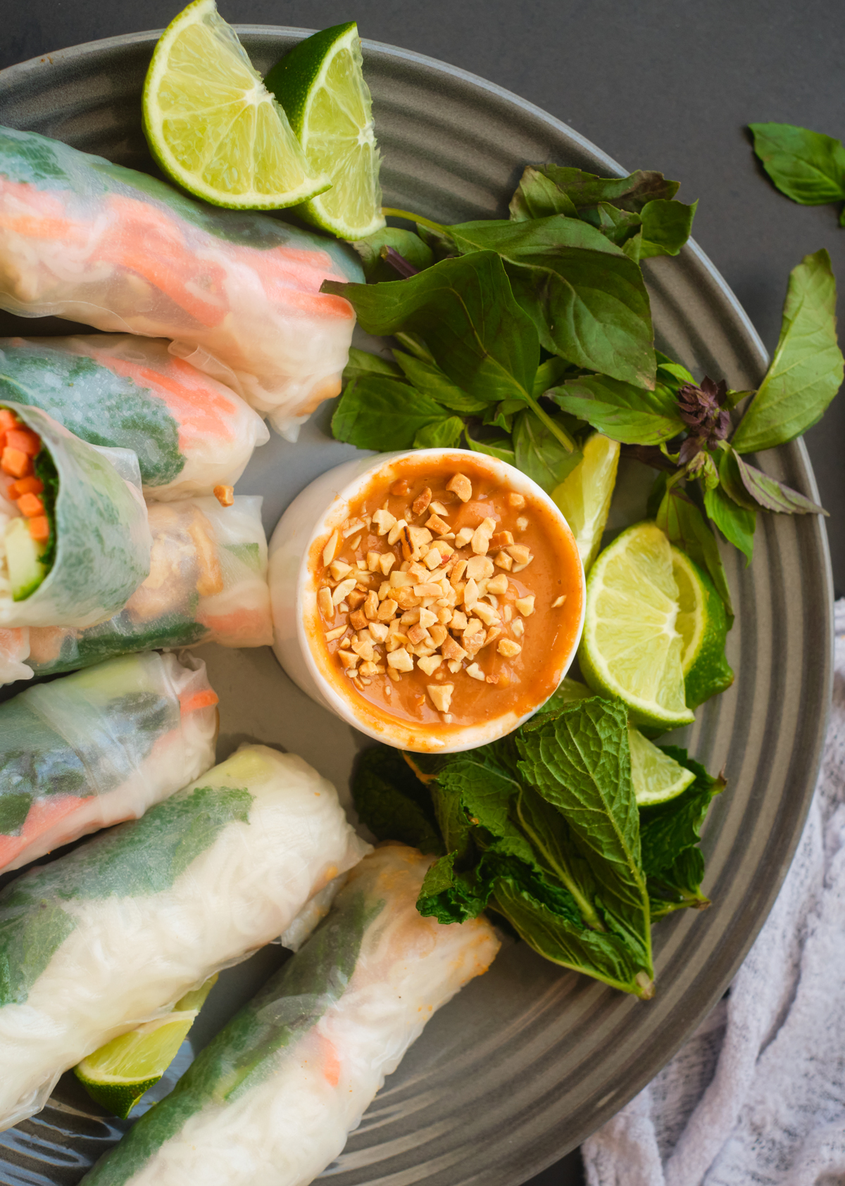 Vietnamese Peanut Sauce with crushed peanuts on top in a single dipping bowl surrounded with Thai Basil, min, lime wedges and lemongrass chicken salad rolls