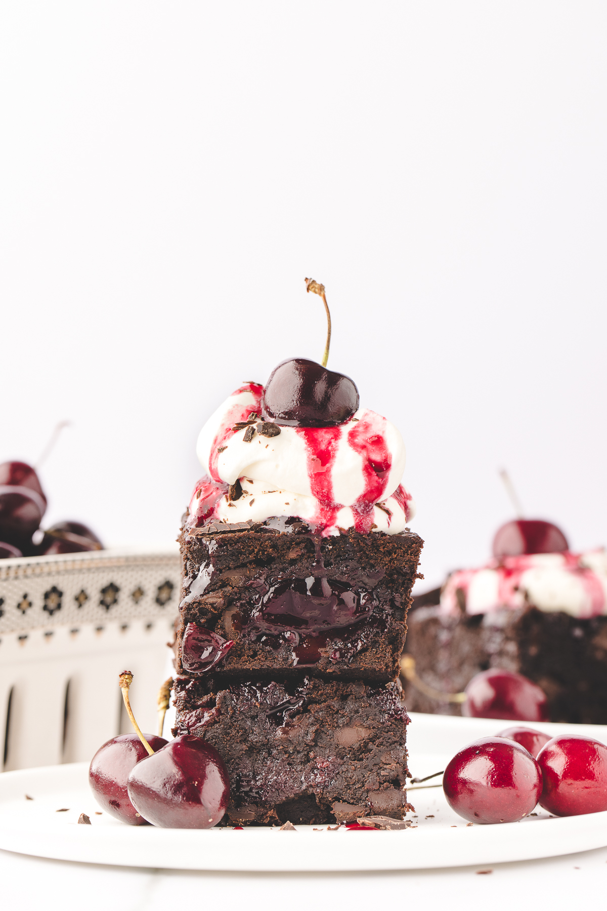 Black Forest Brownies Recipe. Brownies 2 of them stacked with whipped cream on top and a cherry. Brownies in background and cherries in a basket on the left