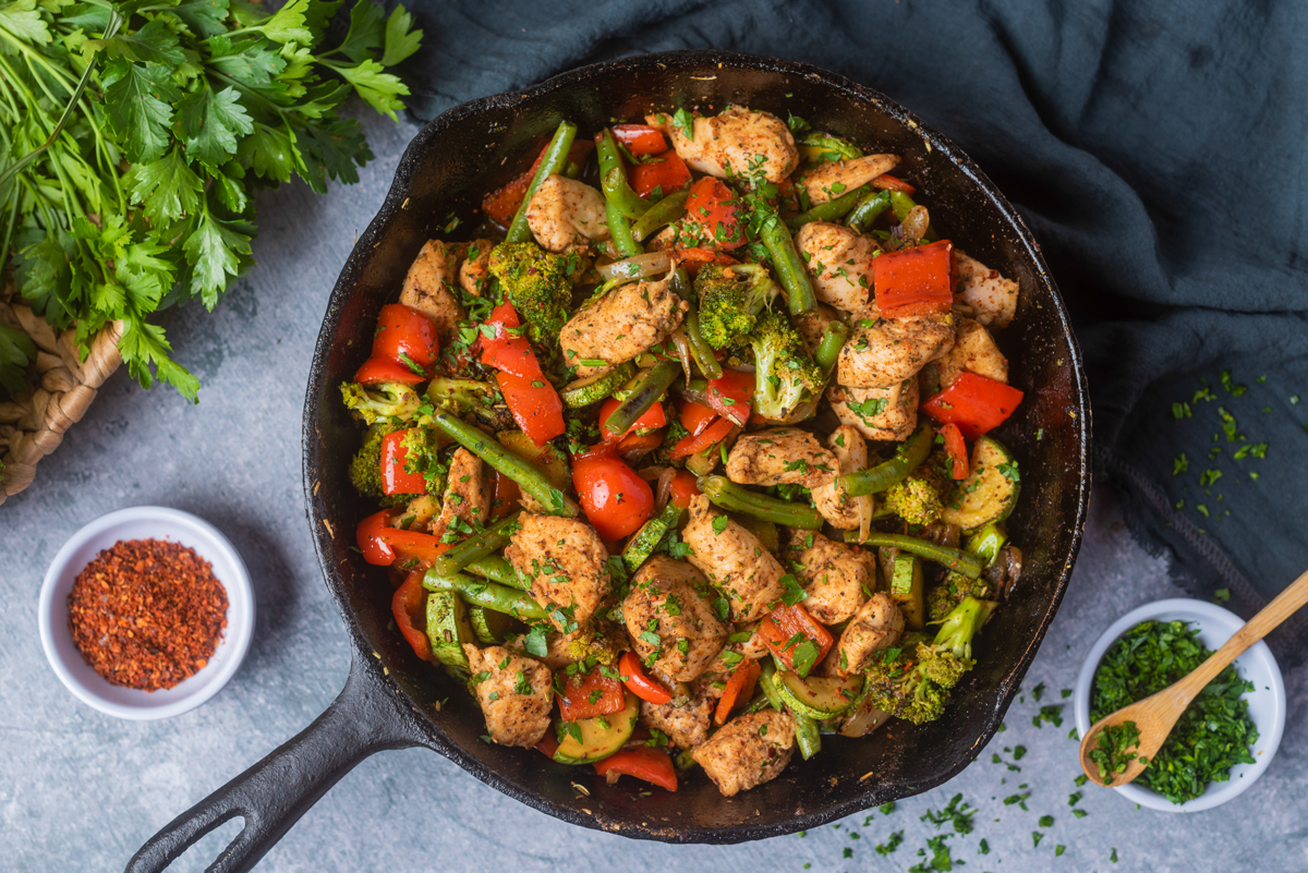 chicken and vegetables skillet Chicken breast bites, red bell peppers, zucchini, green beans and broccoli cooked in a cast iron pan with chopped parsley on top and in the top left corner