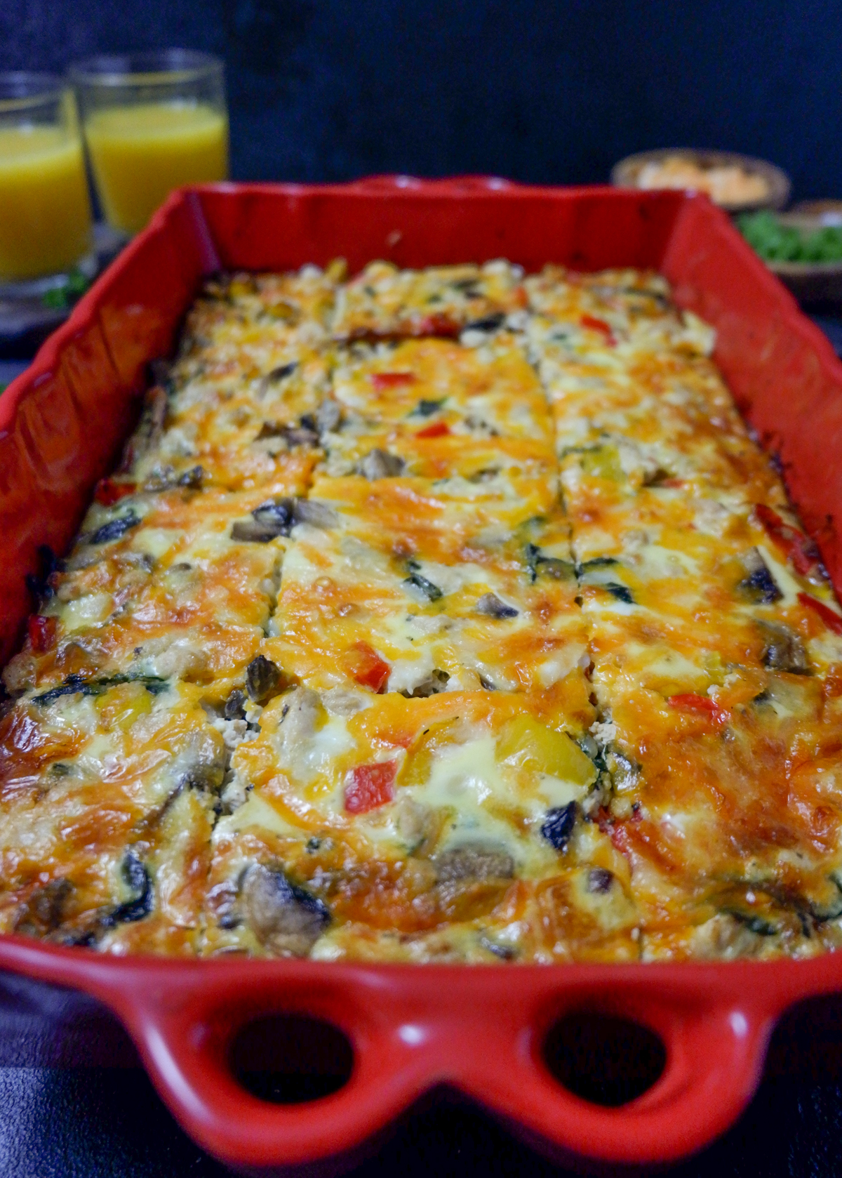 chicken breakfast casserole in a red casserole dish looking from one end to the other portrait style. 2 glasses of oj in the back top left, shredded cheese and parsley in wooden bowls in the top right back