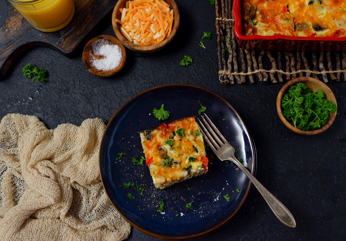 breakfast casserole slice on a blue plate. Shredded cheese, coarse soalt in wood bows in top left, top right casserole dish and parsley in a wood bowl