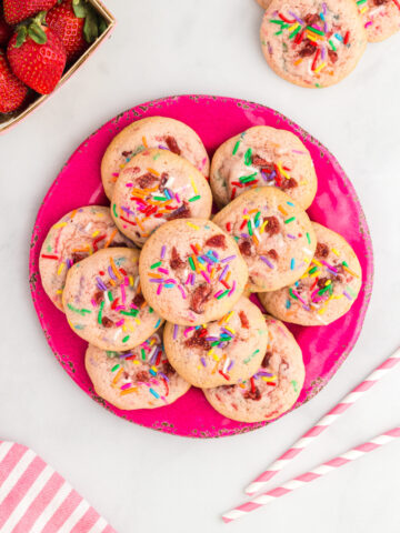 stack of strawberry milkshake cookies on a hot pink plate. 6 cookies on 1st layer, 4 on next layer and one top layer. top left corner strawberries peeking out, top right a cookie, bottom right red & white striped straws, bottom left red and white striped cloth peeking out.