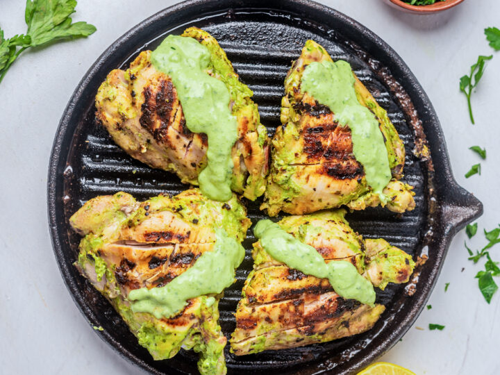 4 green goddess chicken thighs on a grill pan with dressing on top, on a white background, half lemon bottom right, green herbs in a small wooden bowl top right, blue and white striped linen along the top