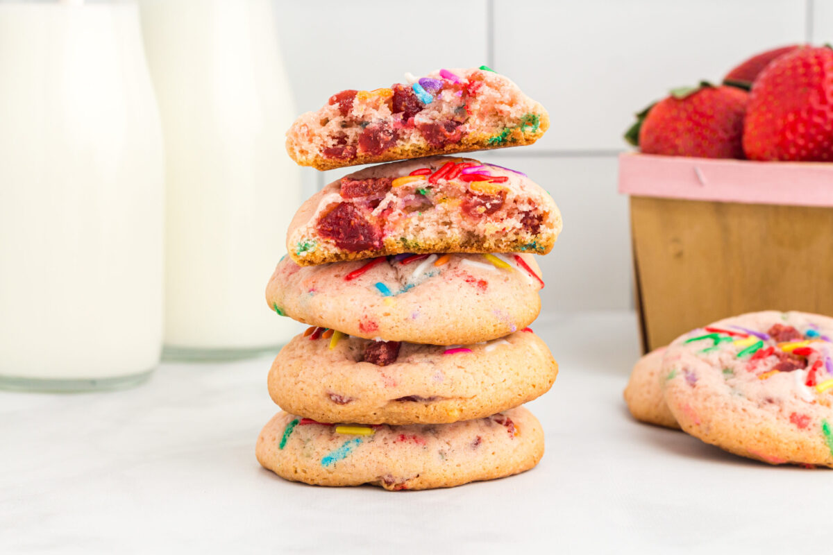 2 jugs of milk on the left, stack of 5 cookies in the middle with the top 2 broken in half, 2 cookies on the right in front of a basket of strawberries, part of the basket showing