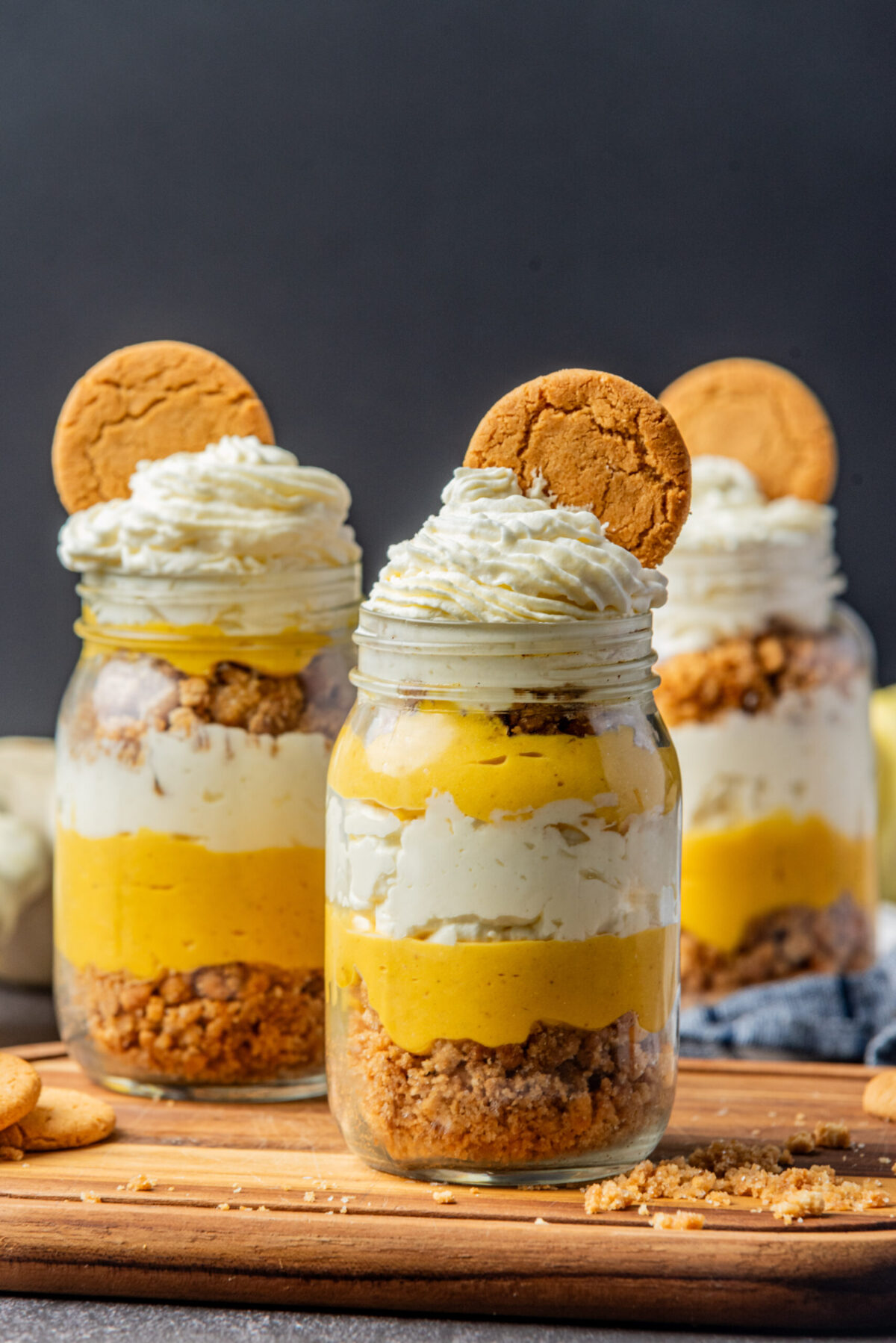 3 mason jars with pumpkin parfait topped with whipping cream/cream cheese mix and a ginger snap cook on a wooden board
