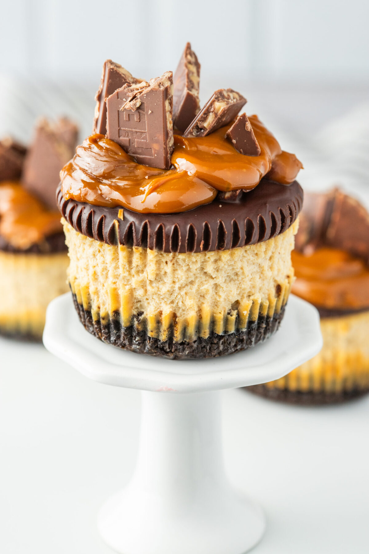 single recipe dulce de leche cheesecake on a mini white cake stand with other cheesecakes behind it.