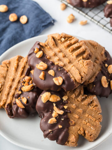 chocolate dipped peanut butter cookies with chopped peanuts on top on a white plate in front, some on a cooling rack to the right. Blue linen on the left.