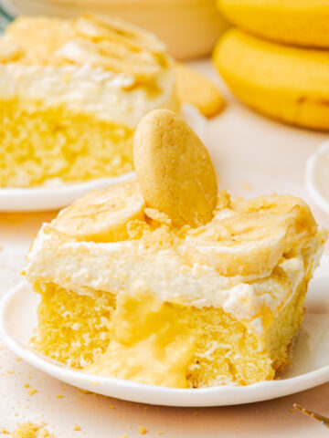 banana pudding poke cake slice on a white plate with a nilla cookie wafer on top and sliced bananas
